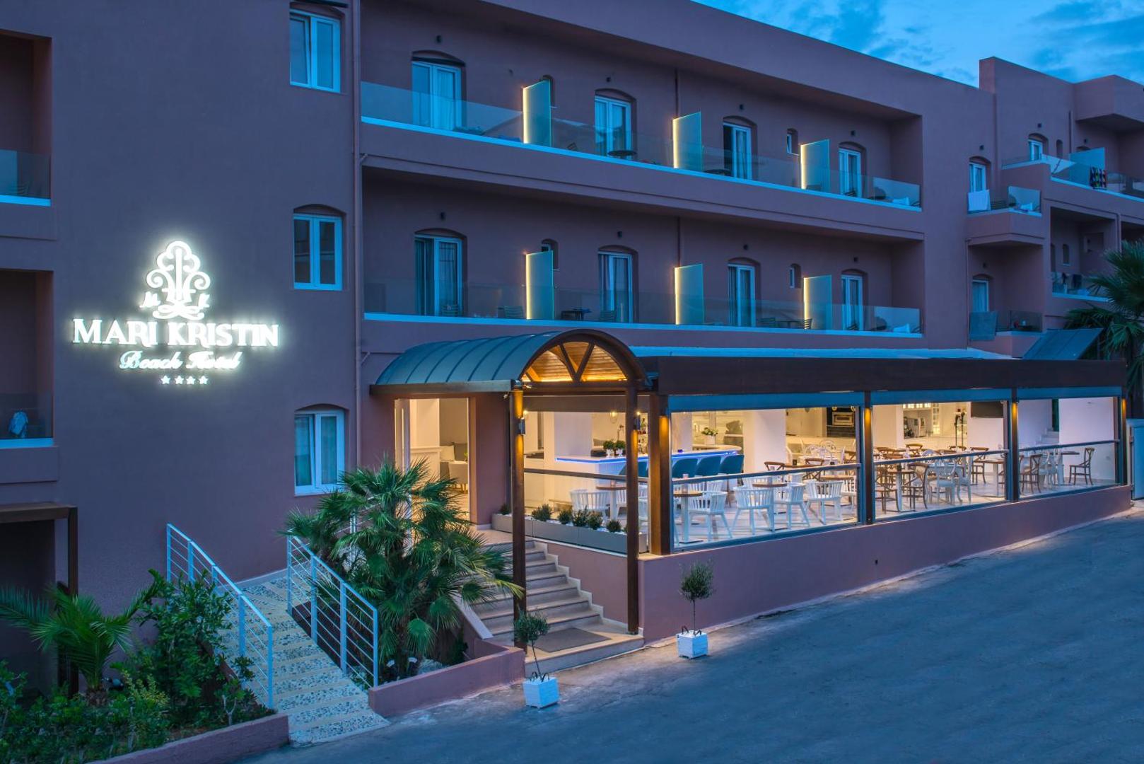 Mari Kristin Beach Hotel fasāde Hersonisā, Krētā — četrzvaigžņu viesnīcas ieeja vakarā ar izgaismotu logo un restorāna terasi