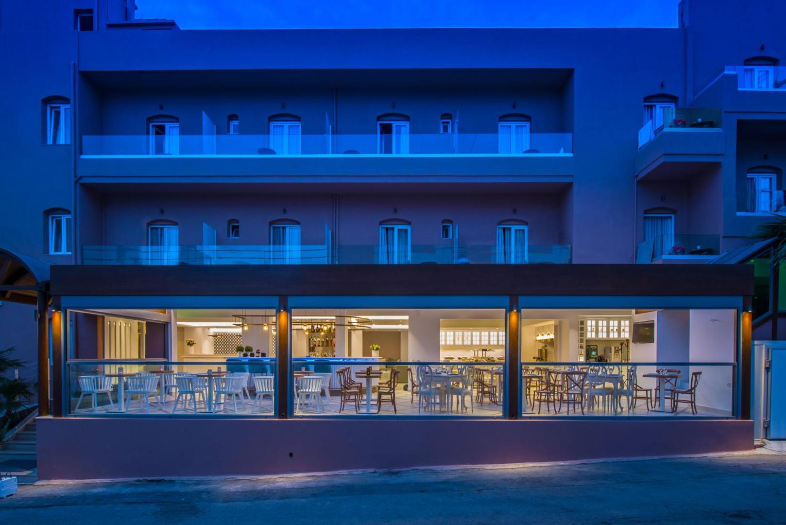 Mari Kristin Beach Hotel restorāna terase vakarā — eleganti balti krēsli un koka galdi zem nojumes ar skatu uz ielu
