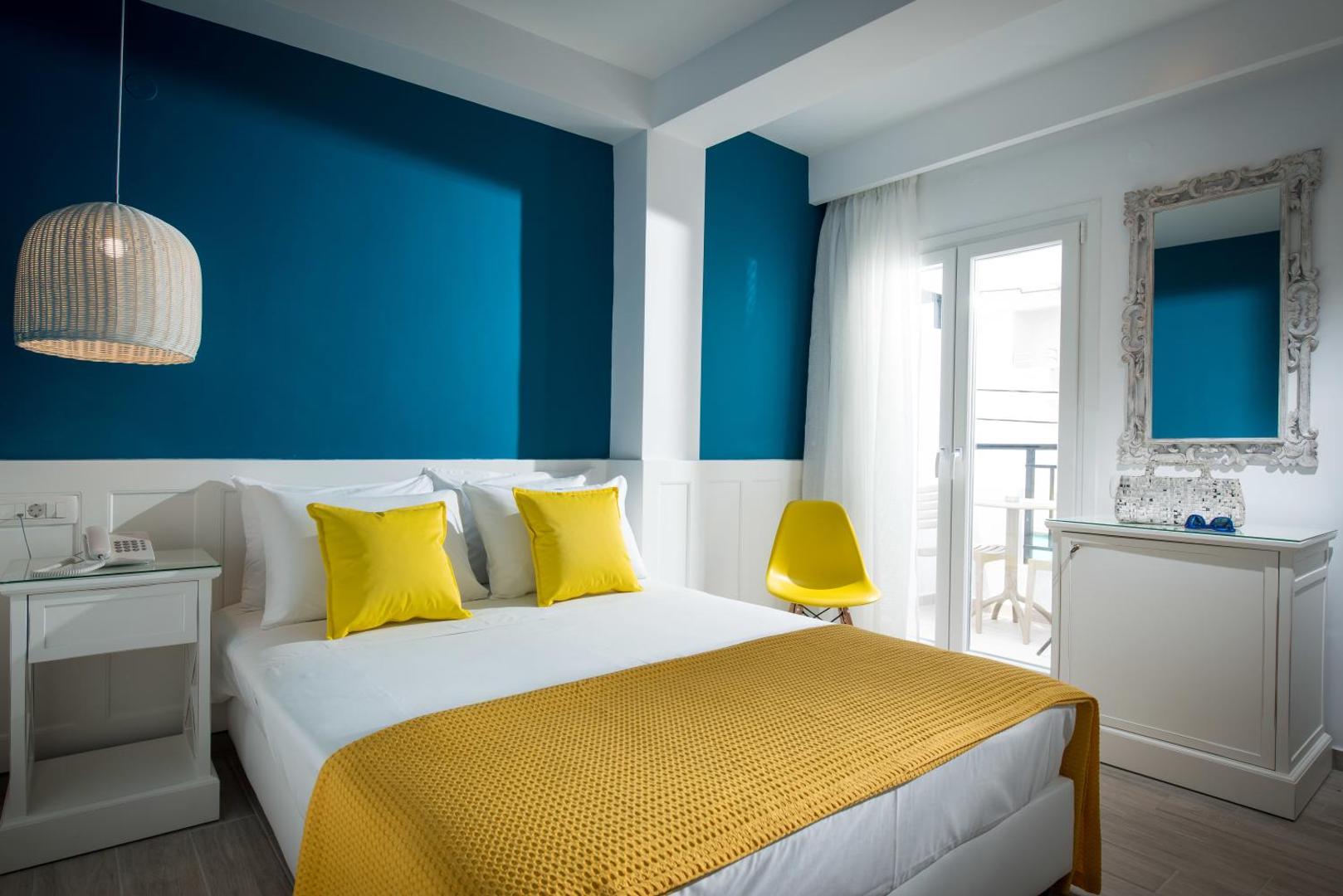 Mari Kristin Beach Hotel moderns numurs ar zilu akcenta sienu, dzelteniem spilveniem un piekļuvi balkonam Krētas salā
