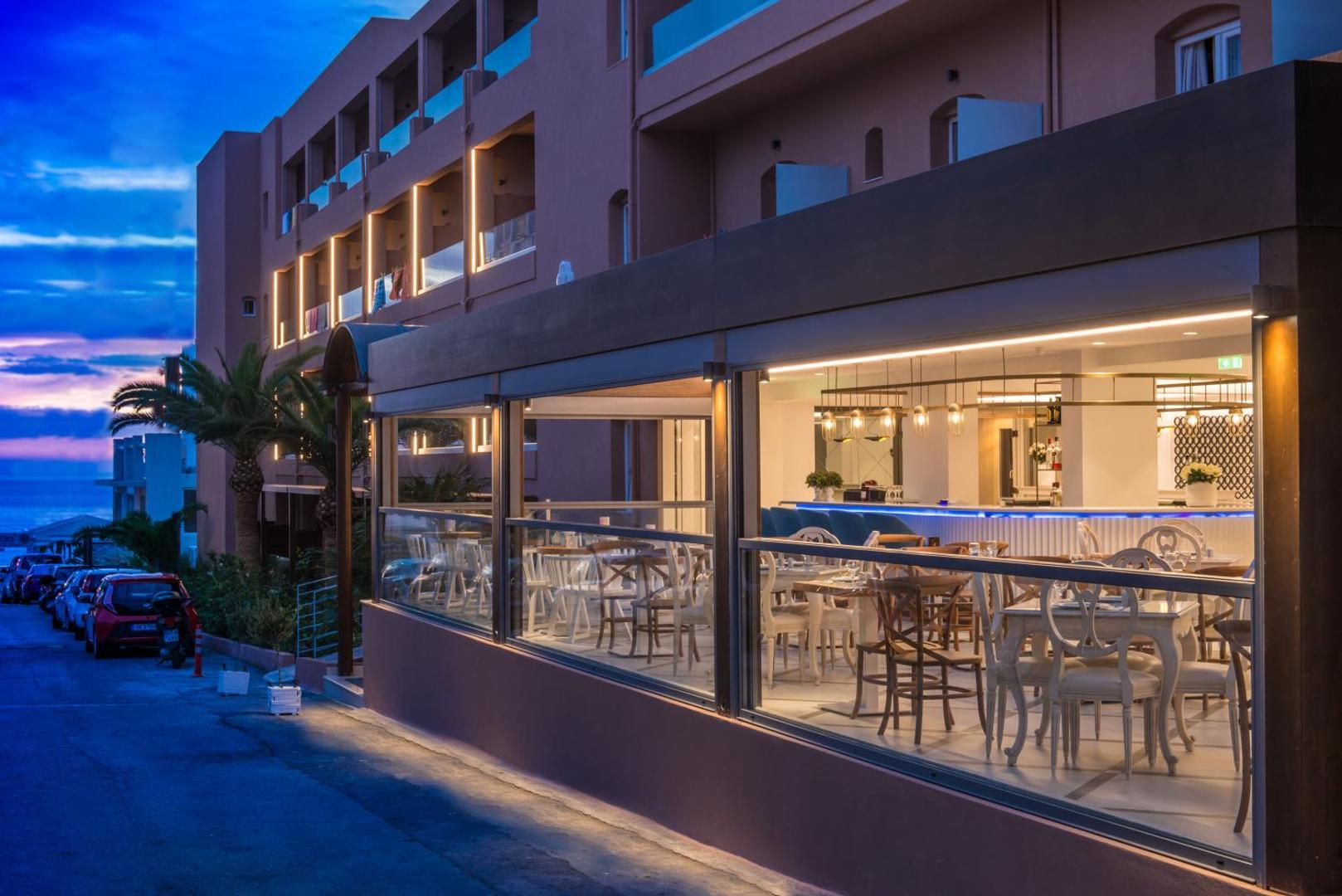 Mari Kristin Beach Hotel bāra un restorāna zona vakarā — moderna stiklota terase ar siltu apgaismojumu un skatu uz jūru tālumā