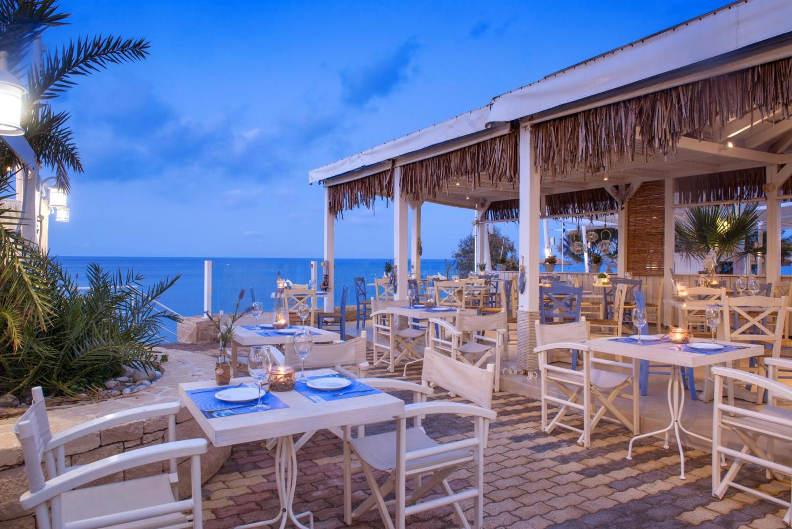 Mari Kristin Beach Hotel terases restorāns ar skatu uz jūru vakarā, balti klāti galdi un grieķu stila interjers Hersonisā