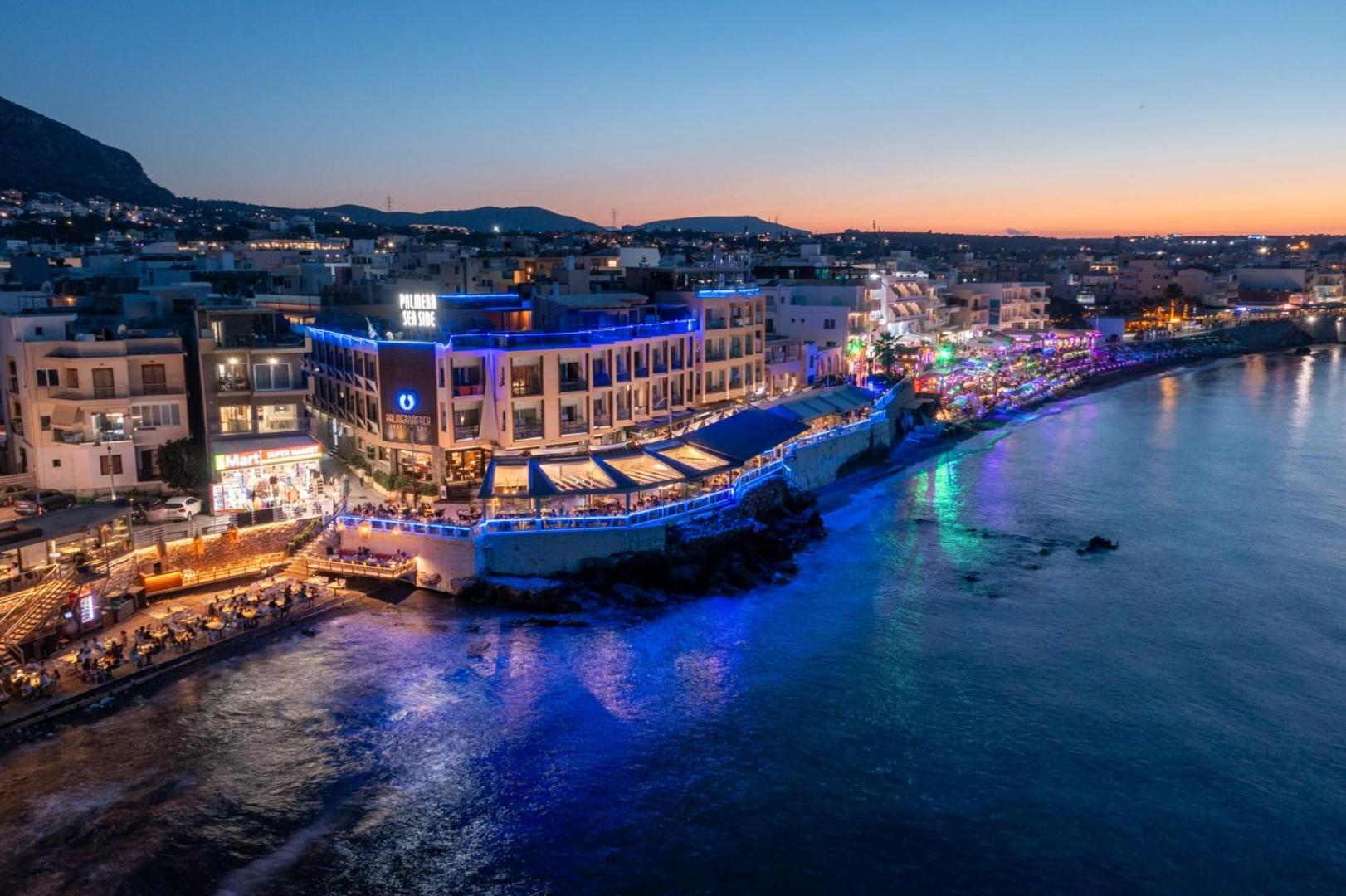 Palmera Beach Hotel & Spa skats no putna lidojuma vakarā Hersonisā, Krētā — izgaismota viesnīcas ēka pie pašas jūras krasta