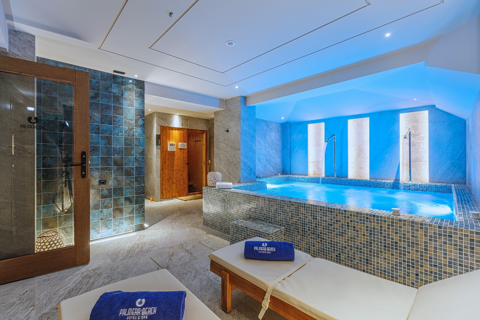 Palmera Beach Hotel & Spa iekštelpu baseins ar zilu apgaismojumu, pirti un atpūtas krēsliem Hersonisā, Krētā — moderna spa zona