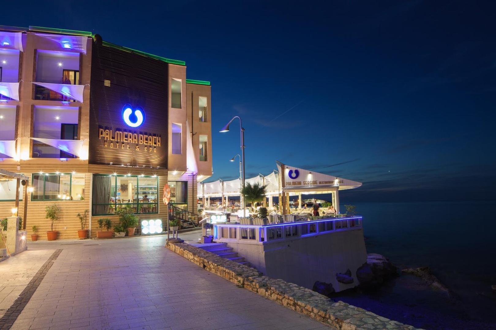 Palmera Beach Hotel & Spa restorāna terase vakarā ar skatu uz jūru — izgaismota fasāde un āra galdiņi piekrastē