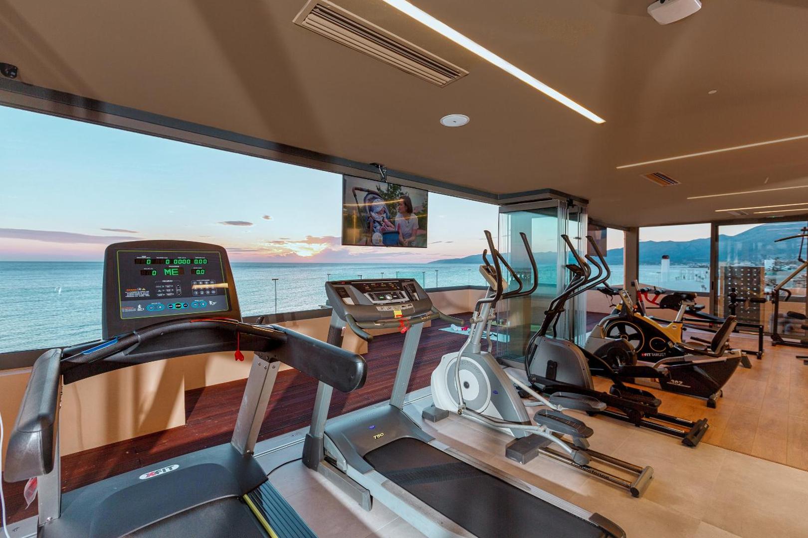 Palmera Beach Hotel & Spa fitness centrs ar skatu uz jūru saulrietā — trenažieri un panorāmas logi