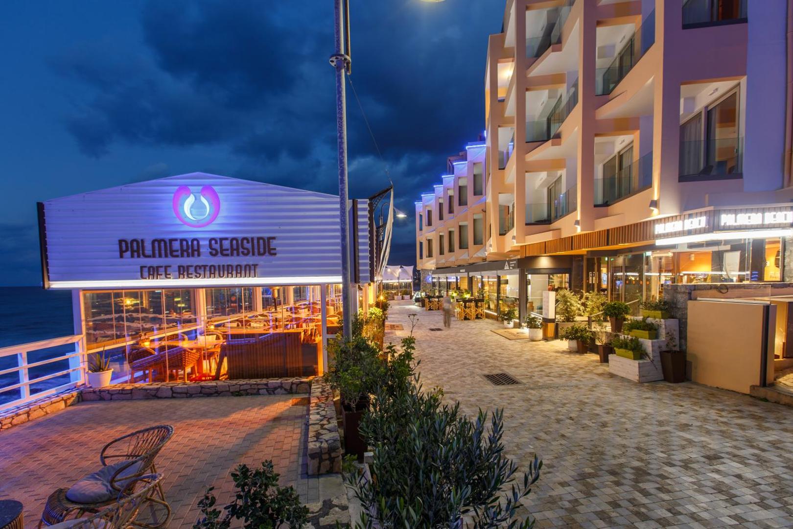 Palmera Seaside restorāna ieeja un viesnīcas fasāde vakarā Hersonisā — moderns apgaismojums un bruģēta gājēju zona pie jūras