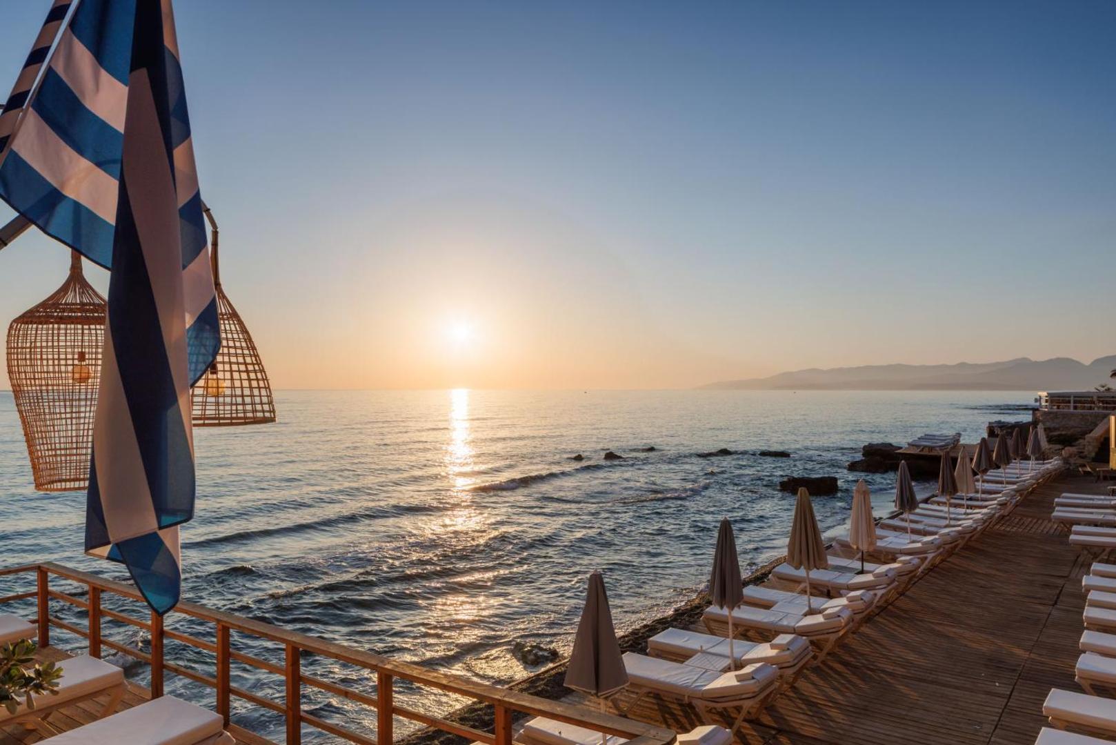 Saulriets Palmera Beach Hotel & Spa pludmales terasē ar sauļošanās krēsliem un Grieķijas karogu fonā Hersonisā