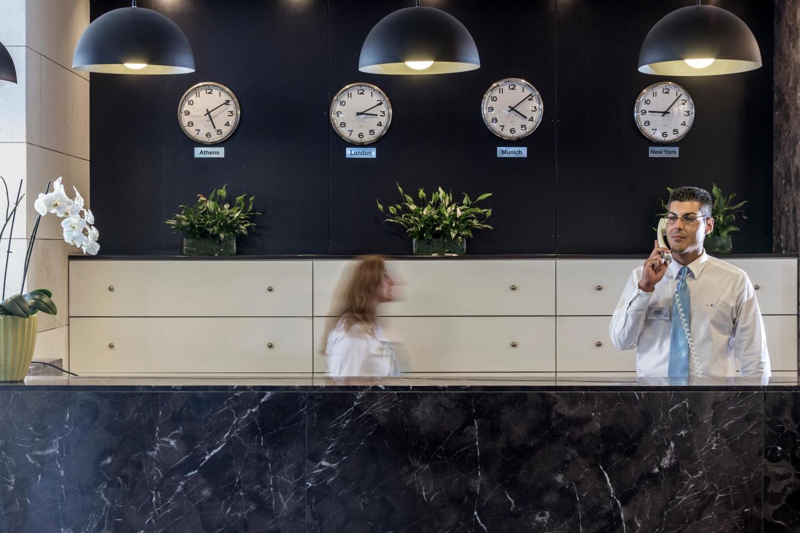 Aeolos Beach Resort viesnīcas recepcijas zona Korfu salā ar tumša marmora leti, sienas pulksteņiem un personālu darbā