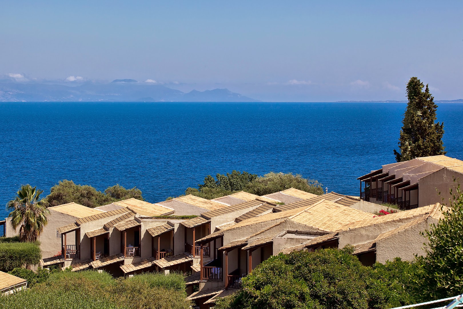 Aeolos Beach Resort bungalo rindu mājas ar terasēm Korfu piekrastē — fonā zilā Jonijas jūra un kalnu silueti