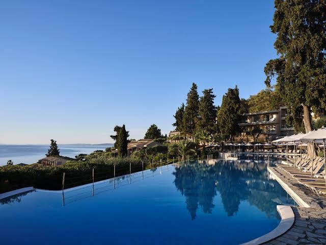 Aeolos Beach Resort baseins ar pārplūdes malu, sauļošanās krēsliem un saulessargiem Korfu piekrastē, ieskauts zaļos kokos un kalnos.