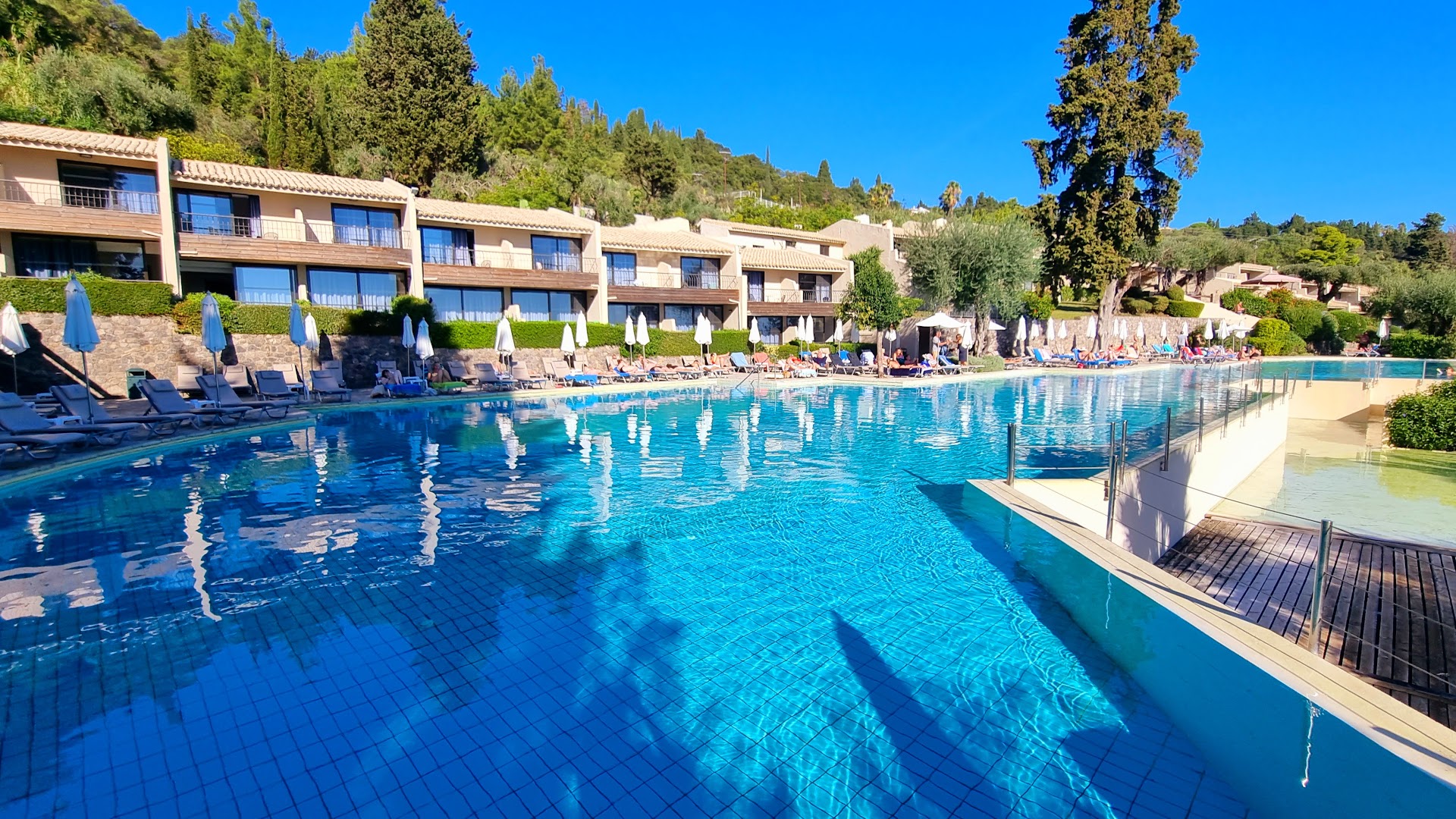Aeolos Beach Resort galvenais āra baseins Korfu salā ar skatu uz viesnīcas korpusiem, saulessargiem un apkārtējo zaļo kalnu ainavu