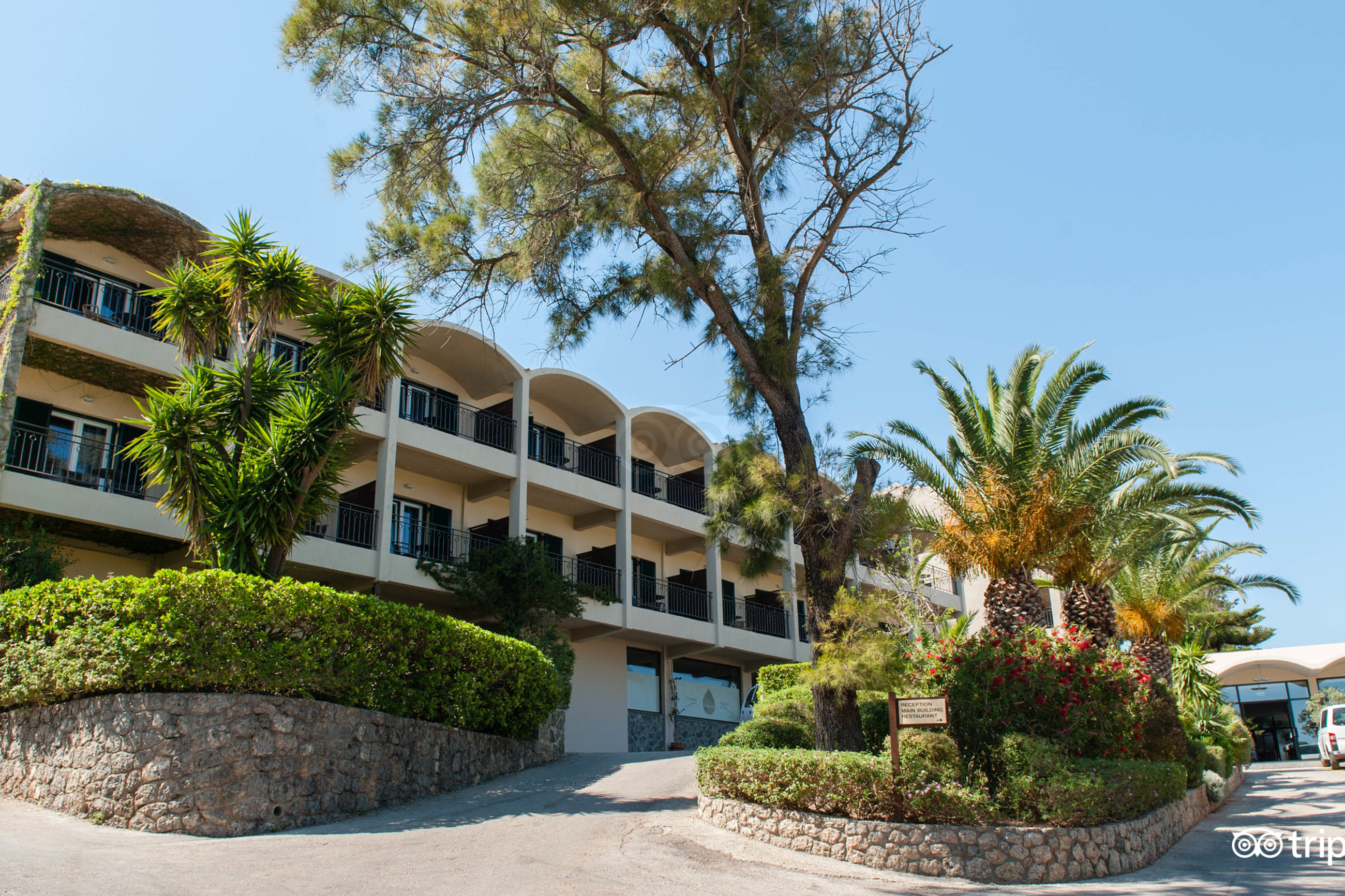 Aeolos Beach Resort galvenā ēka ar balkoniem un eksotiskiem augiem, palmām un priedēm Korfu — ieeja viesnīcas recepcijā un restorānā