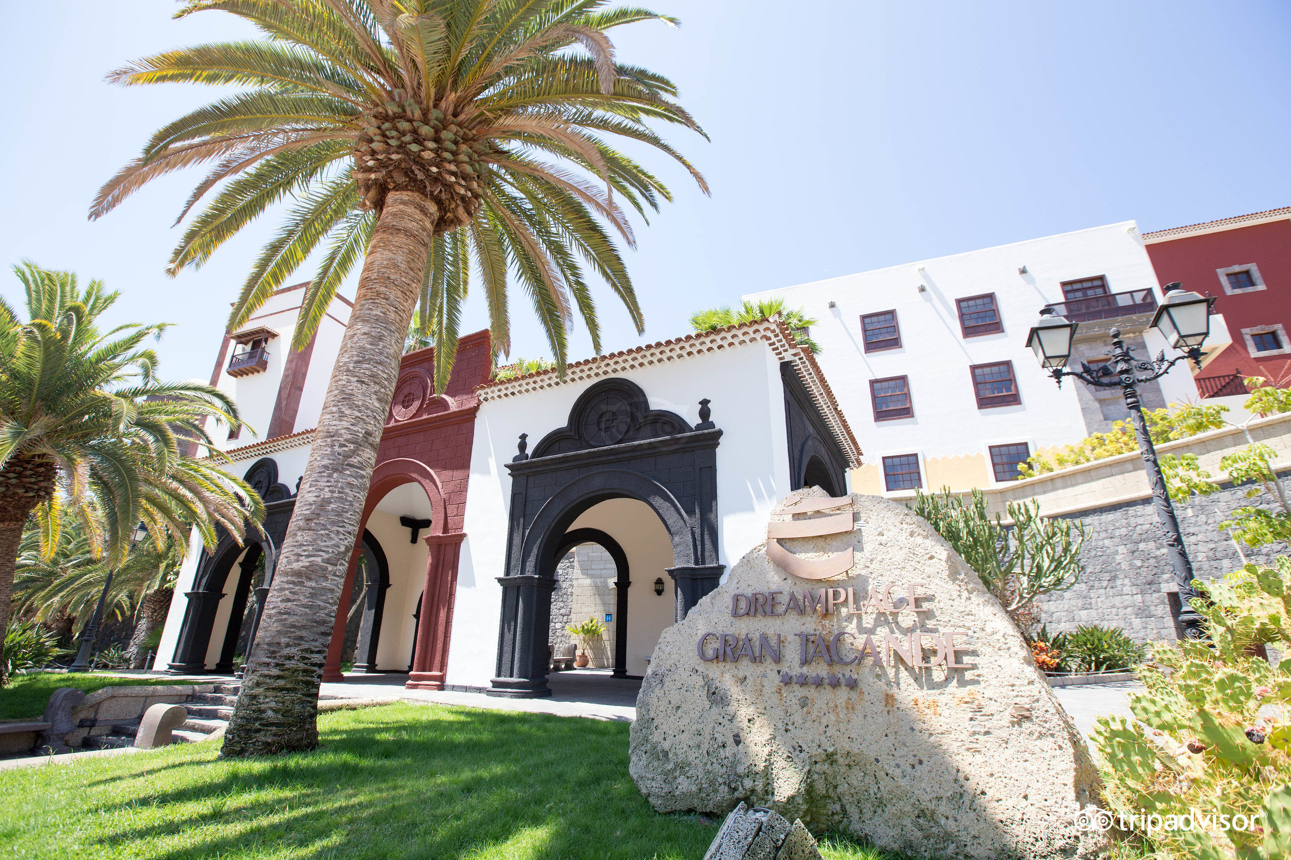 Viesnīcas Gran Tacande Wellness & Relax ieeja Tenerifē ar akmens zīmi, palmām un tradicionālo Kanāriju salu arhitektūru saulainā dienā.