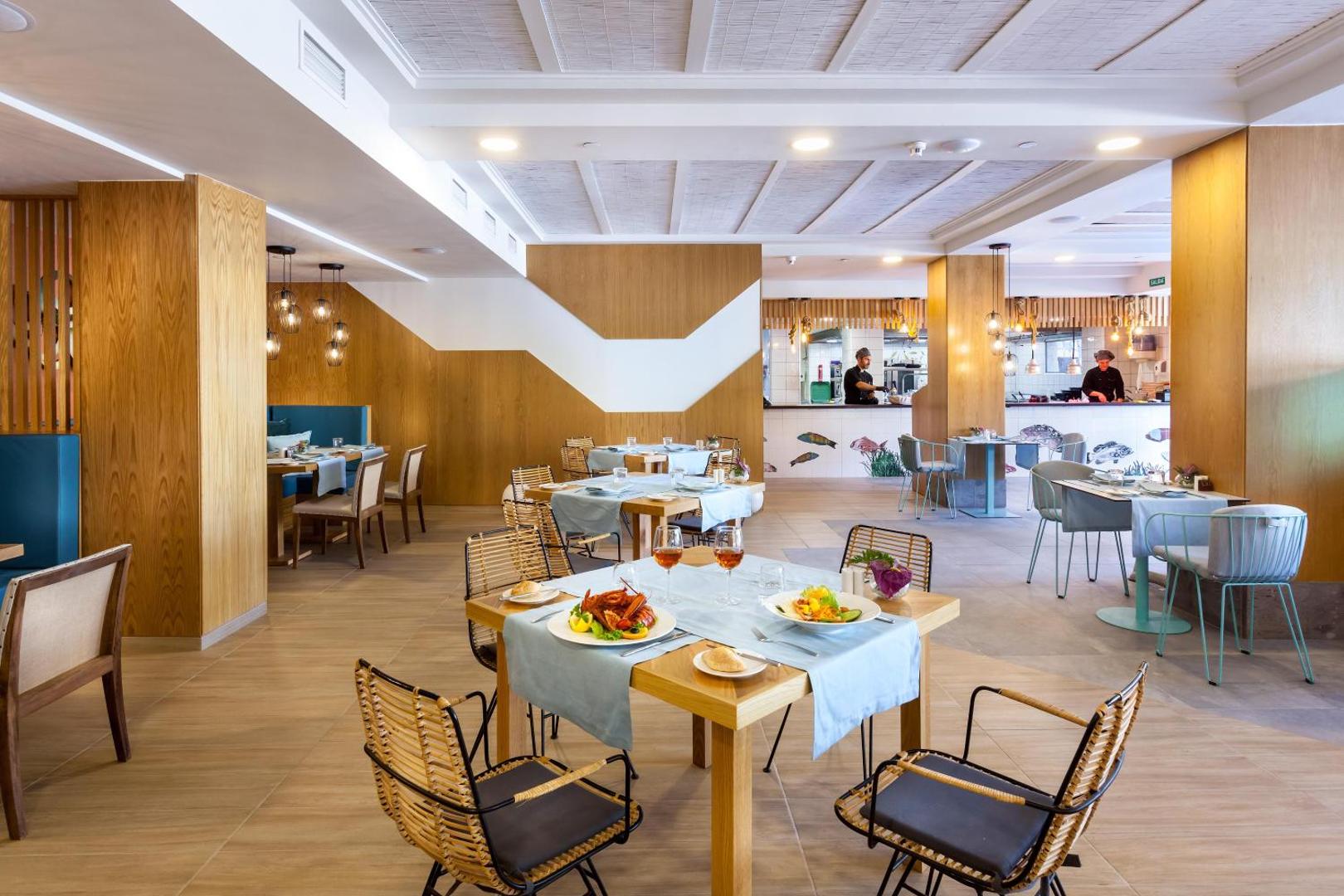 Gran Tacande Wellness & Relax restorāns ar modernu koka interjeru, klātiem galdiem un atvērto virtuvi fonā