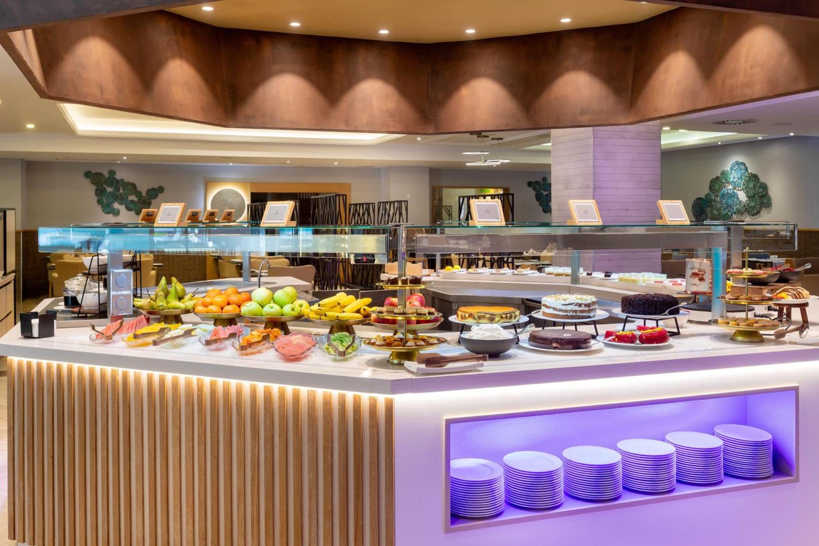 Gran Tacande Wellness & Relax restorāna desertu un augļu bufete ar kūkām, svaigiem augļiem un modernu violetu apgaismojumu.