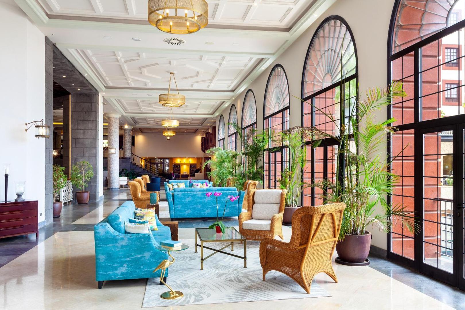 Gran Tacande Wellness & Relax viesnīcas plašais vestibils ar augstiem griestiem, lieliem logiem, ziliem dīvāniem un elegantiem pītiem krēsliem.