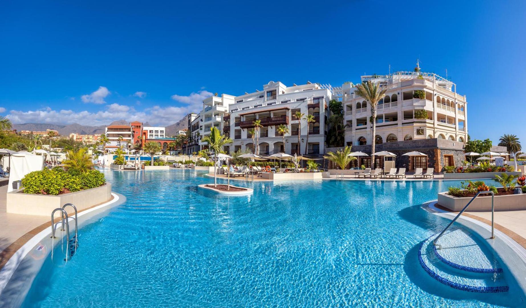 Gran Tacande Wellness & Relax plašais āra baseins ar gaišām ēkām, palmām un kalnu fonu saulainā dienā Tenerifē