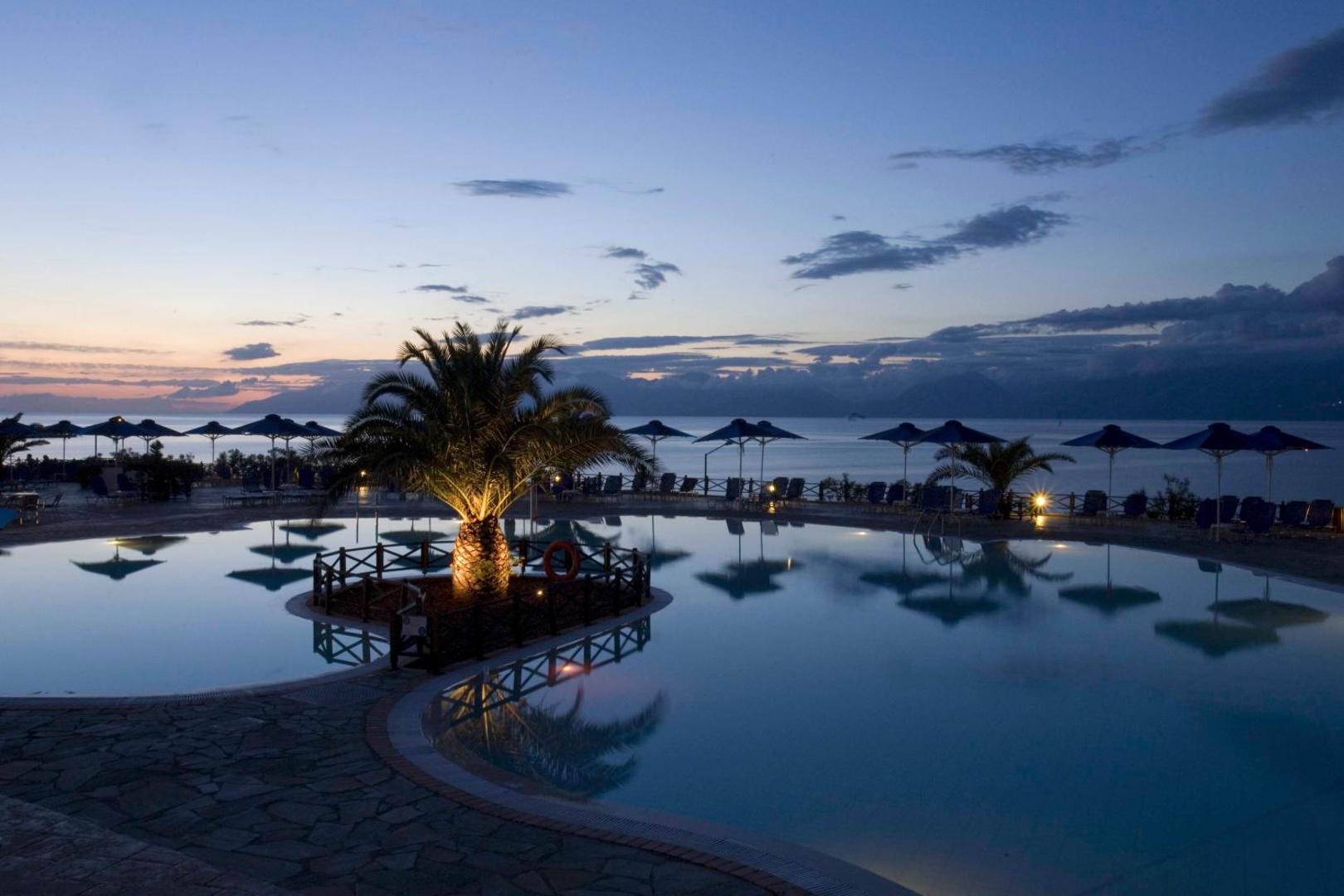Mareblue Beach Resort baseins krēslas stundā ar izgaismotām palmām un skatu uz jūru Korfu piekrastē