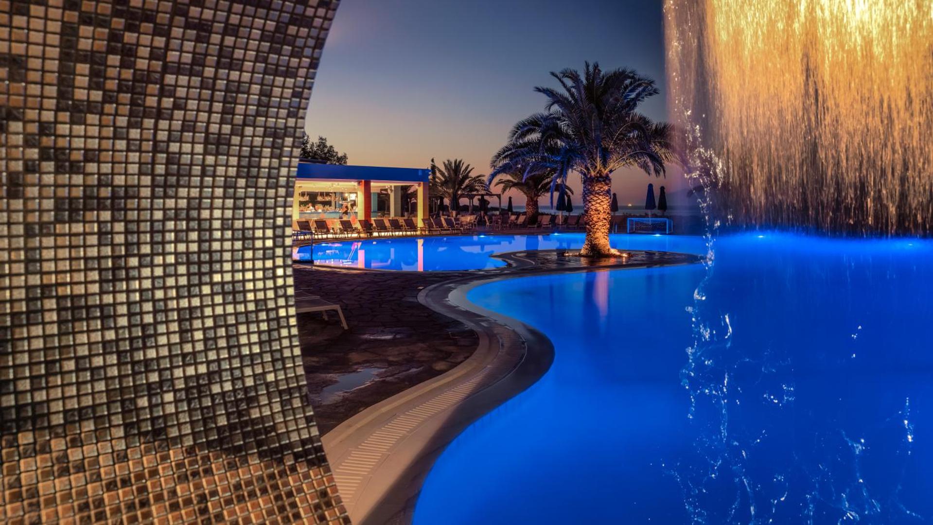 Mareblue Beach Resort āra baseins vakarā ar zilu apgaismojumu un dekoratīvu ūdens kritumu Korfu salā, Grieķijā