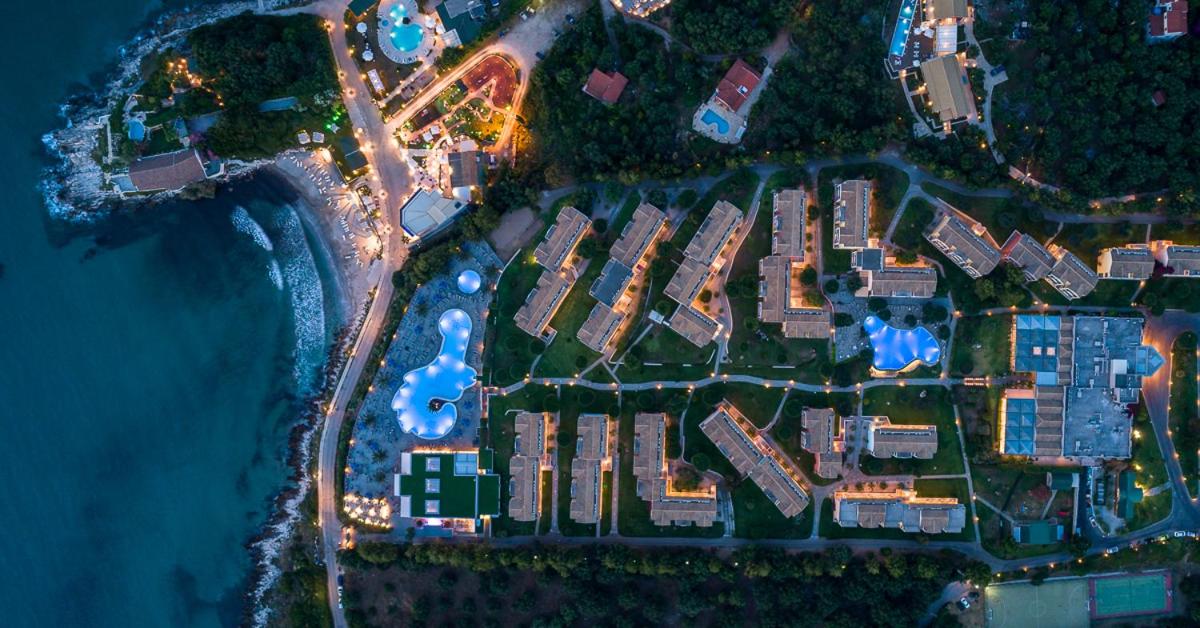 Mareblue Beach Resort skats no putna lidojuma vakarā — izgaismoti baseini, viesnīcas korpusi un piekrastes līnija Korfu ziemeļos