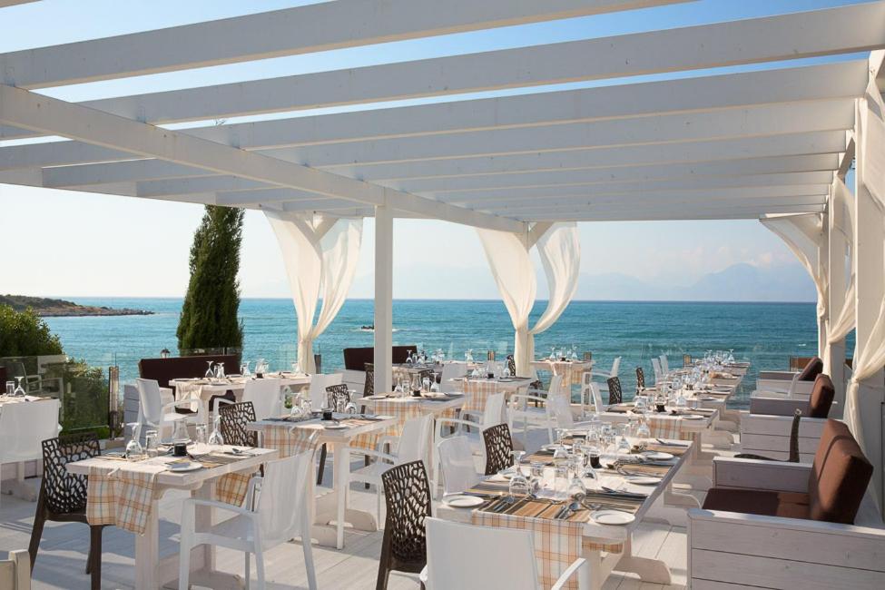Mareblue Beach Resort āra terases restorāns ar skatu uz Jonijas jūru Korfu salā — balti klāti galdi zem koka pergolas.