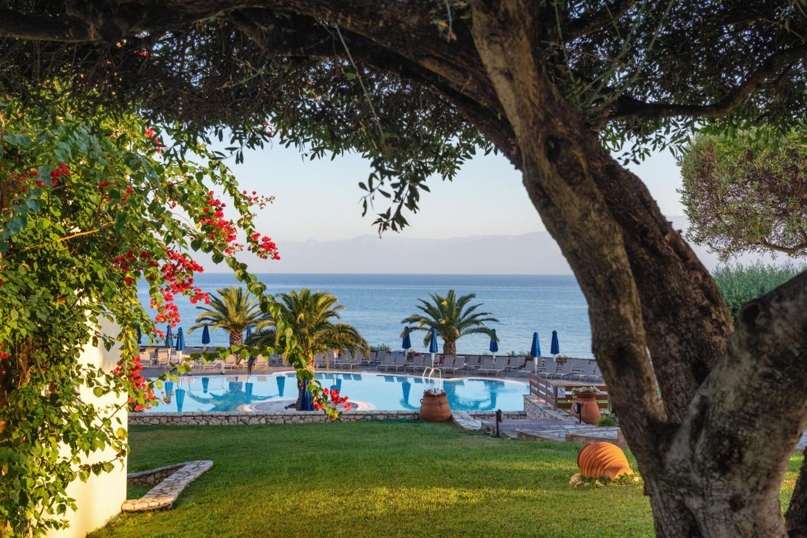 Skats uz Mareblue Beach Resort baseinu un Jonijas jūru caur koku zariem un ziedošiem krūmiem Korfu piekrastē