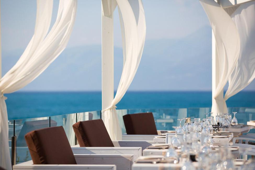 Tuvplāns Mareblue Beach Resort restorāna galdiem pie stikla margām ar skatu uz zilo jūru un kalniem tālumā.