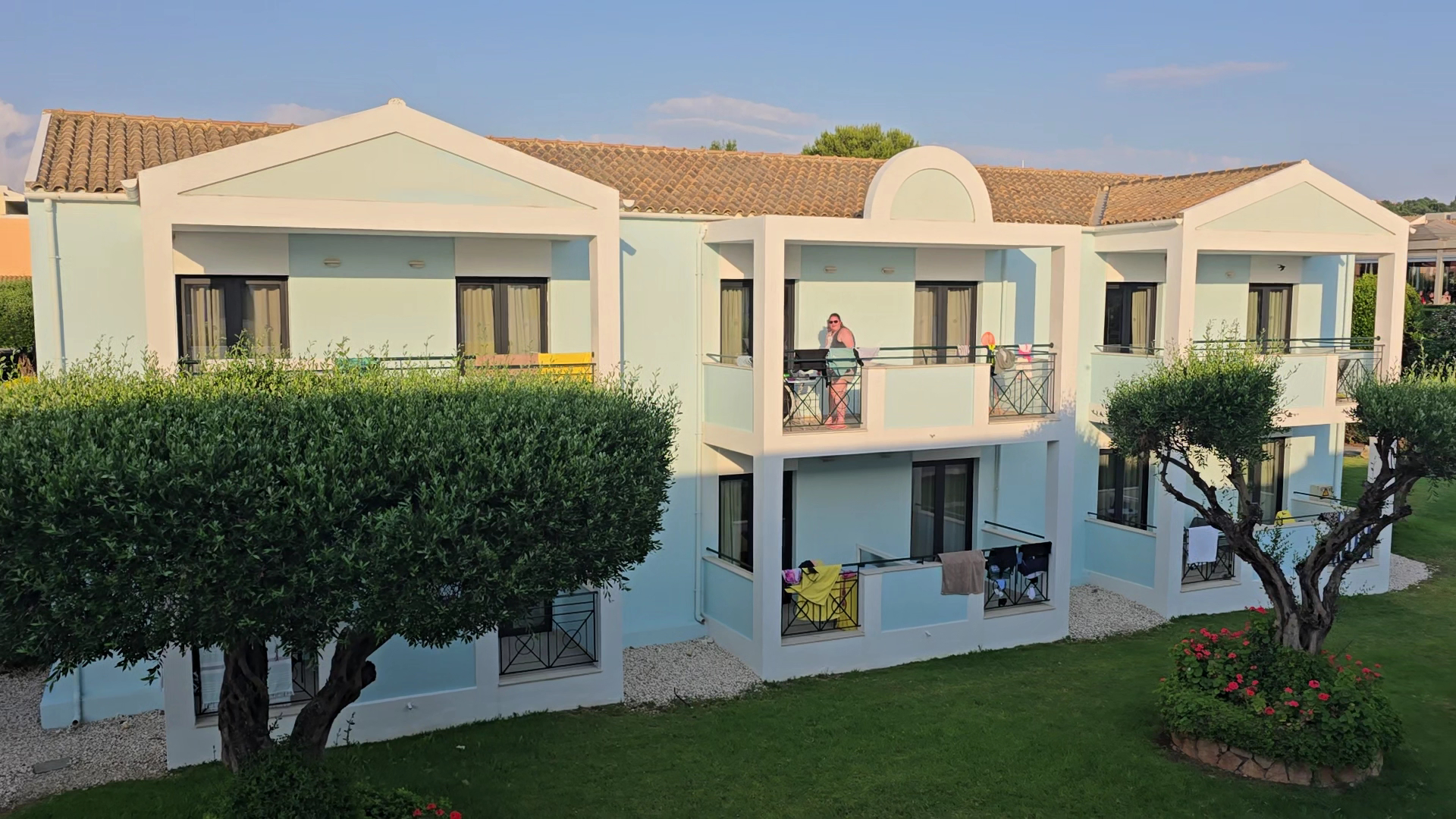 Mareblue Beach Resort gaiši zilie numuru korpusi ar balkoniem, olīvkokiem un koptu mauriņu viesnīcas teritorijā