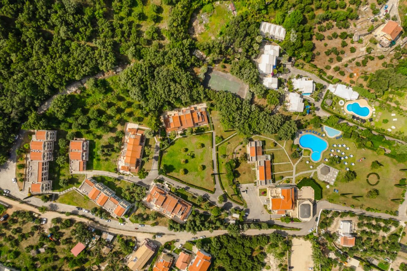 Century Resort Hotel teritorijas skats no putna lidojuma Korfu — redzami baseini, tenisa korts, dārzi un viesnīcas korpusi meža ielokā