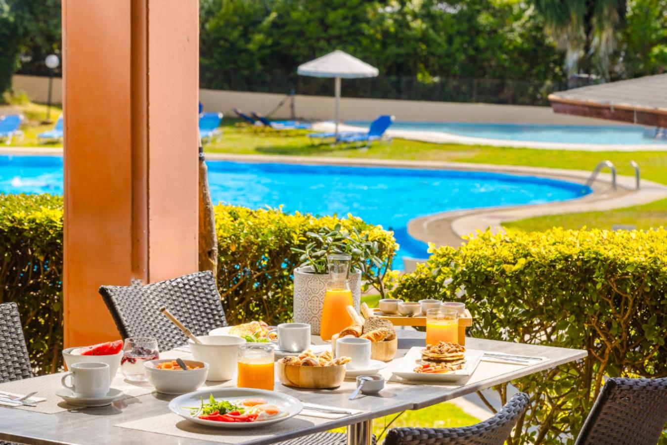 Century Resort brokastu galds uz terases ar skatu uz zilu āra baseinu un dārzu — svaigi augļi un sulas