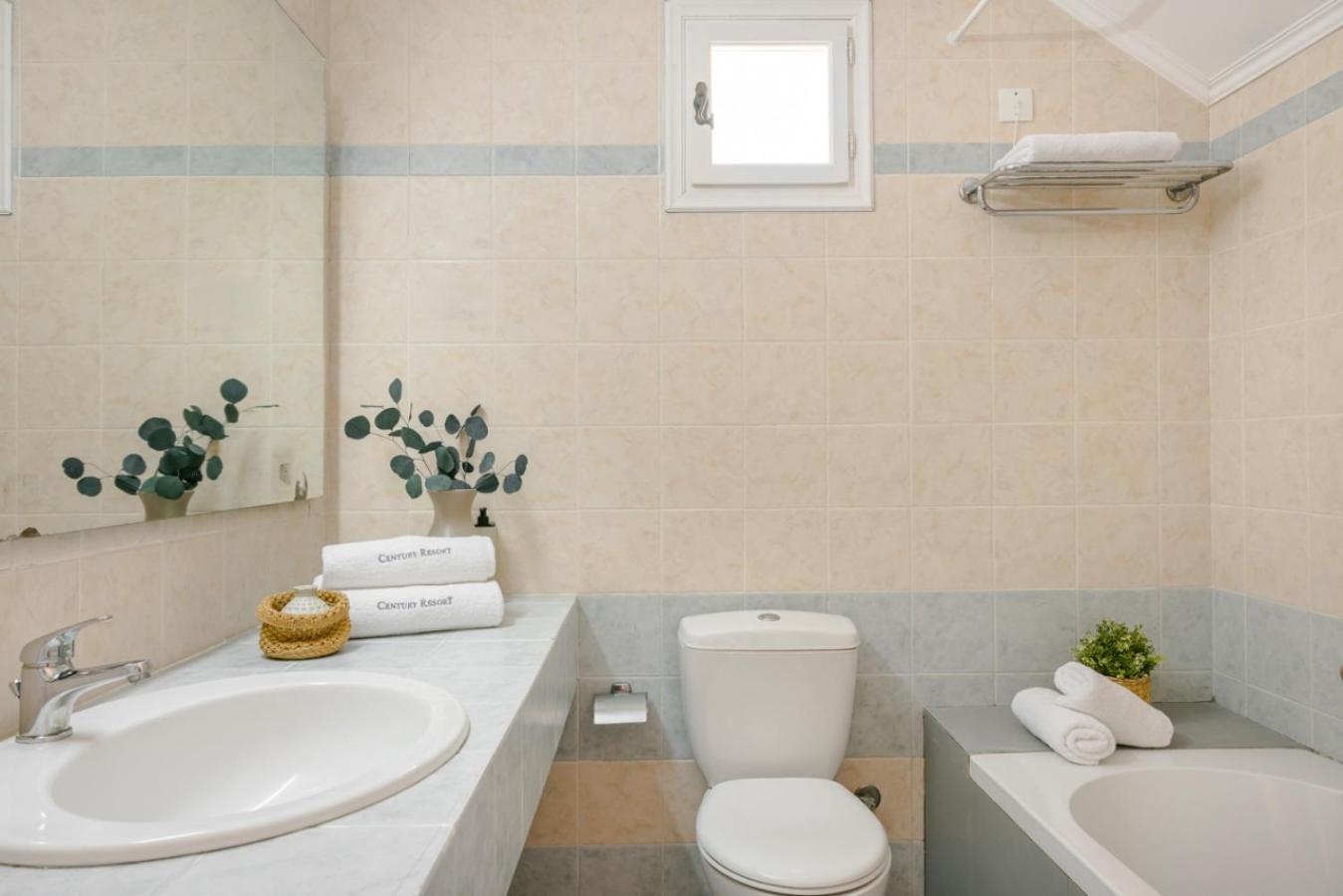 Century Resort Hotel gaišs vannas istabas interjers ar baltu izlietni, spoguli un vannu — tīri dvieļi un dekoratīvi augi.