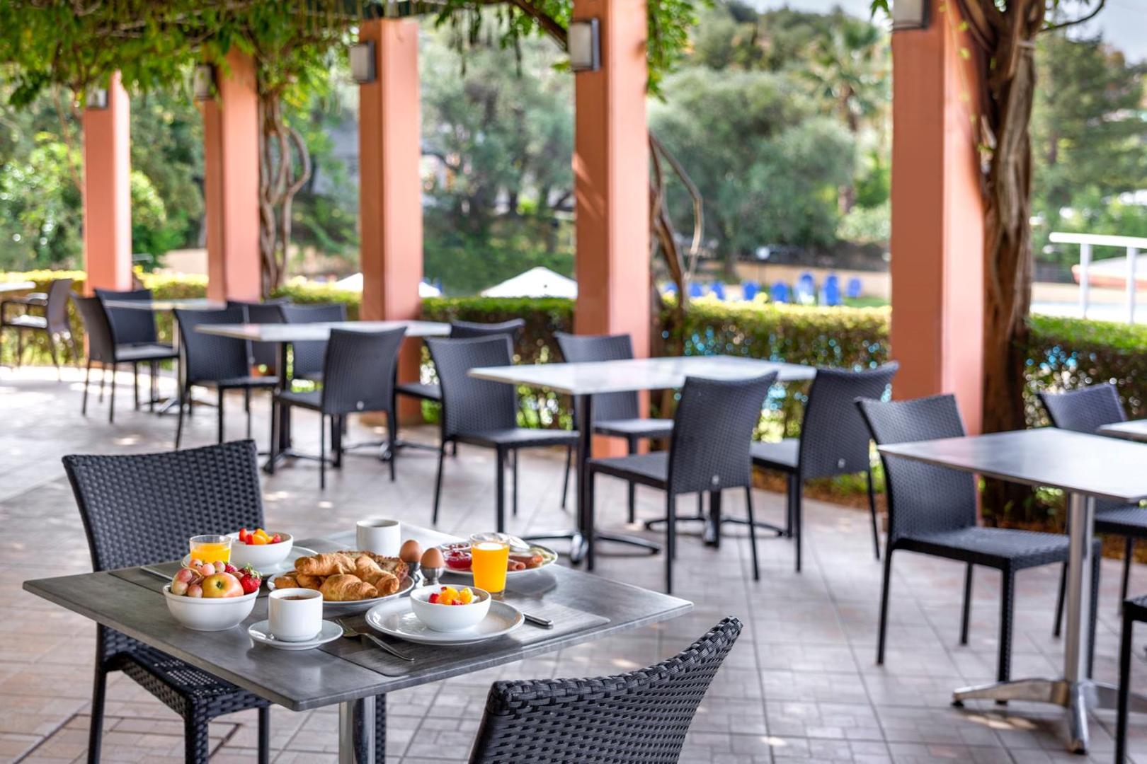 Century Resort segtā āra terase ar brokastu galdiem, pītiem krēsliem un skatu uz zaļo viesnīcas teritoriju