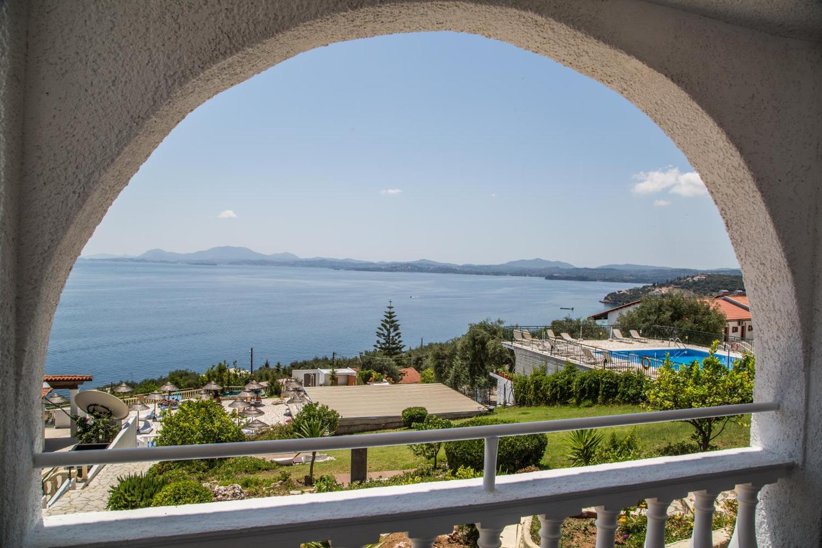 Skats no Pantokrator Hotel balkona arkas uz Jonijas jūru un Korfu piekrasti — zils ūdens un kalnu ainava