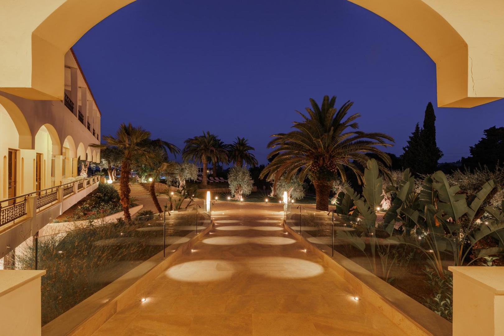 Paradise Corfu viesnīcas ieeja un terase vakarā ar dekoratīvo apgaismojumu, palmām un tumši zilām debesīm fonā