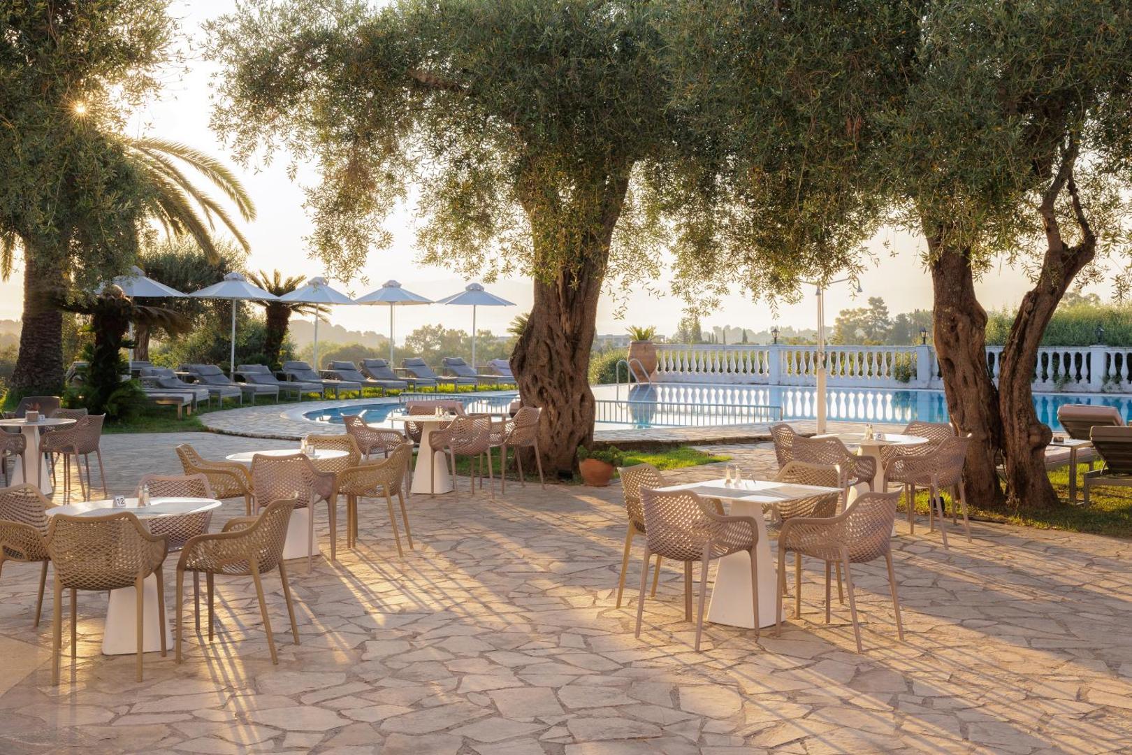 Paradise Corfu āra terase ar galdiņiem un krēsliem olīvkoku ēnā blakus baseinam, skats saulrietā Korfu salā