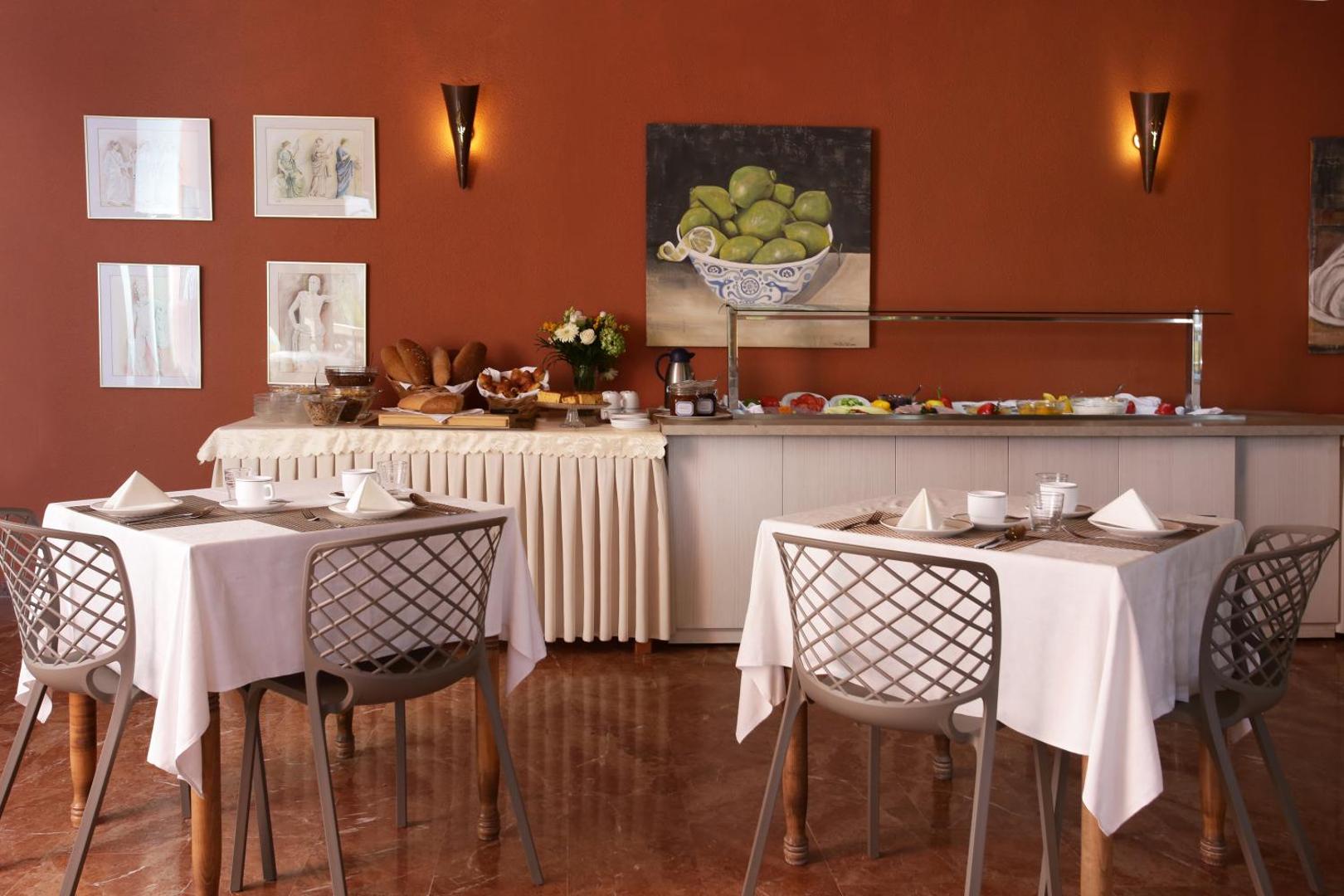Paradise Corfu restorāna brokastu bufetes zona ar klātiem galdiem, gleznām pie sienām un siltu apgaismojumu