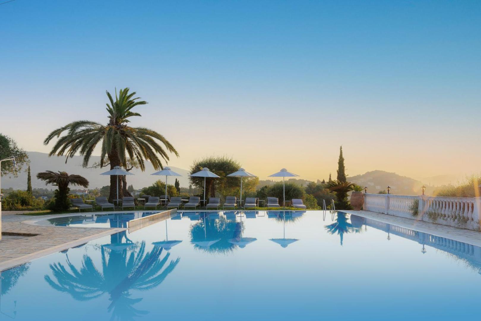 Paradise Corfu viesnīcas āra baseins saulrietā ar palmām un saulessargiem Korfu salā, Grieķijā — skats uz kalniem un dārzu