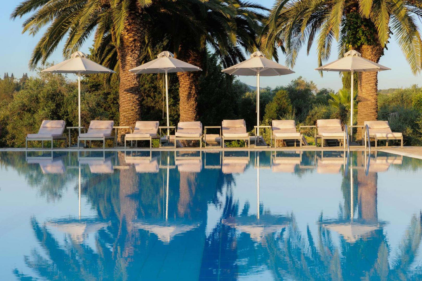 Rinda ar saulessargiem un atpūtas krēsliem pie Paradise Corfu baseina, fonā zaļojošas palmas un dārzs Korfu salā