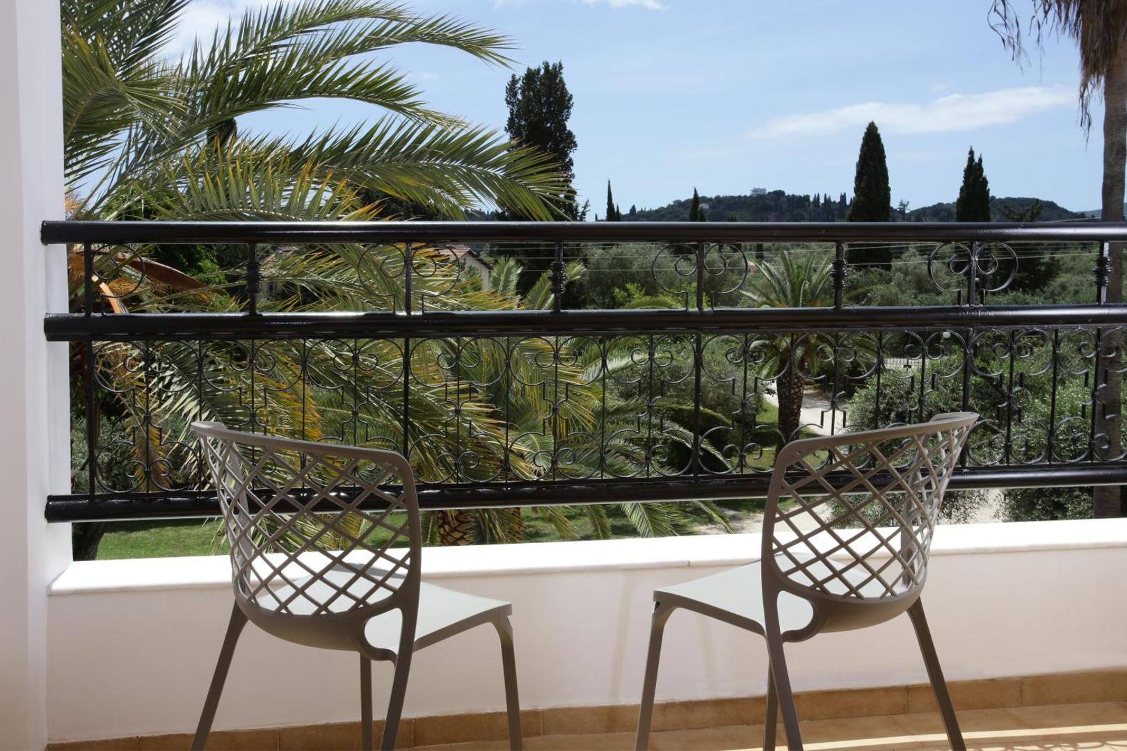 Skats no Paradise Corfu viesnīcas balkona uz palmu dārzu un Korfu kalniem, priekšplānā divi krēsli.
