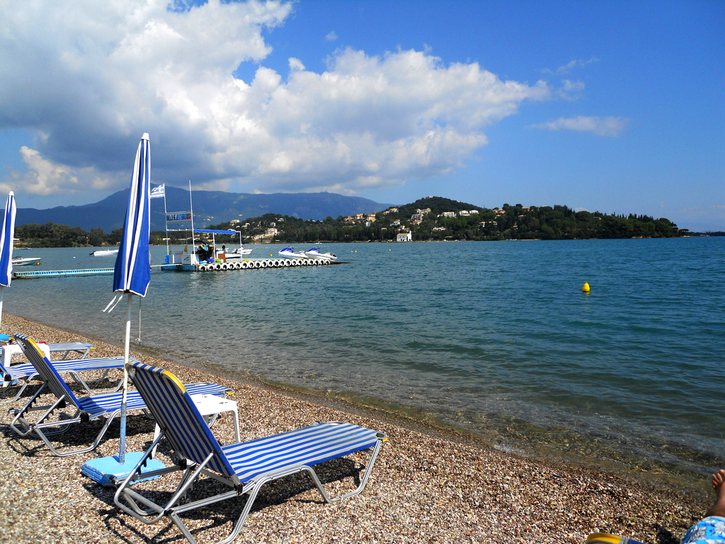 Paradise Corfu oļu pludmale ar zili svītrainiem sauļošanās krēsliem un saulessargiem, skats uz Jonijas jūru un kalniem tālumā