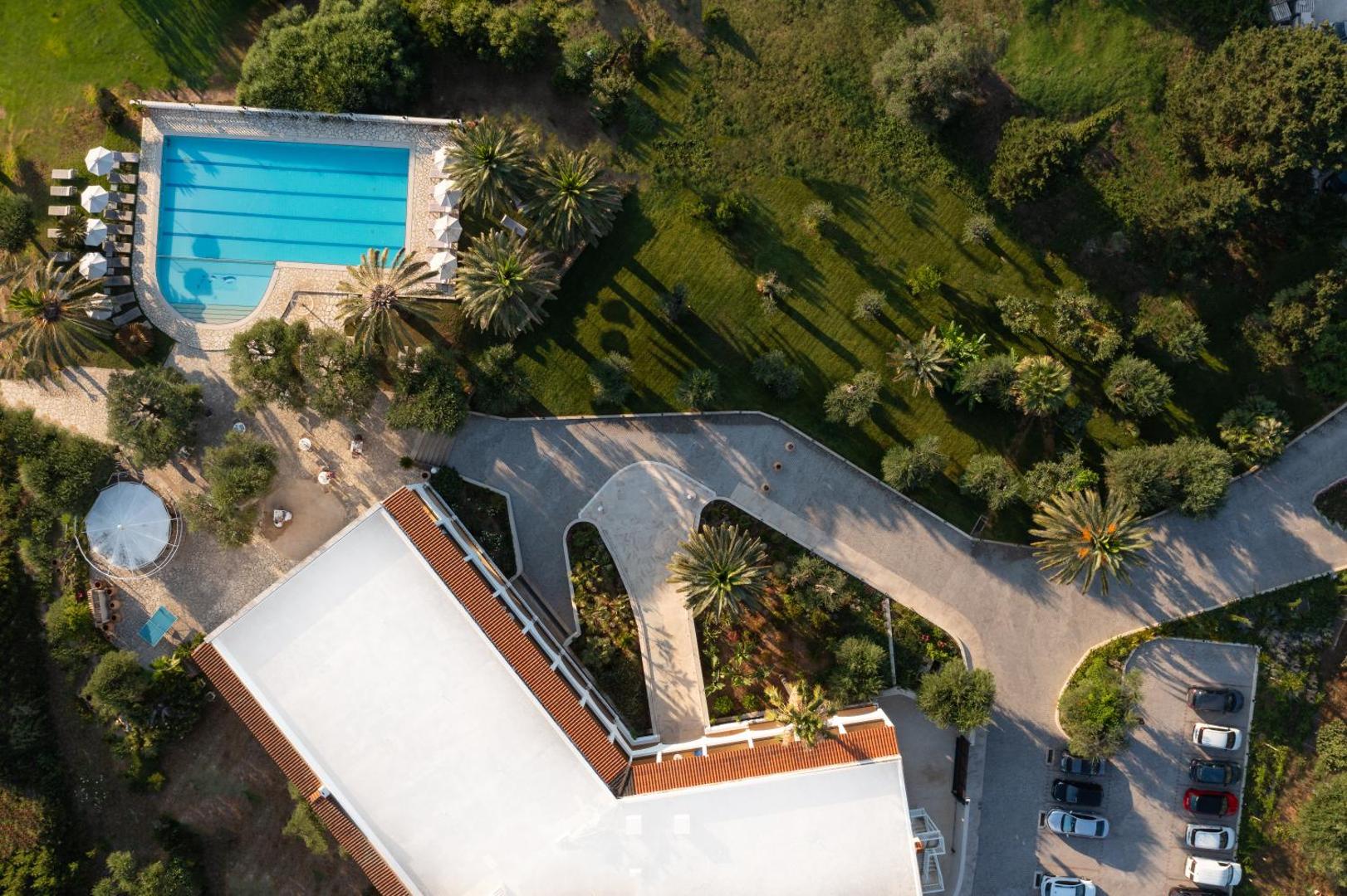 Paradise Corfu viesnīcas teritorijas skats no putna lidojuma — liels āra baseins, sauļošanās zona, palmas un autostāvvieta