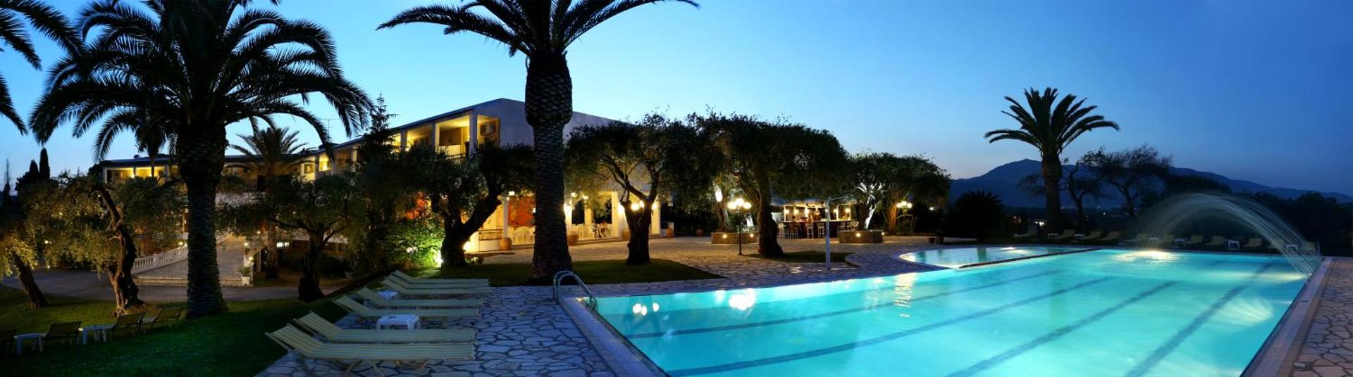Paradise Corfu viesnīcas teritorija vakarā ar izgaismotu baseinu, palmām un galdiņiem pie galvenās ēkas Korfu salā