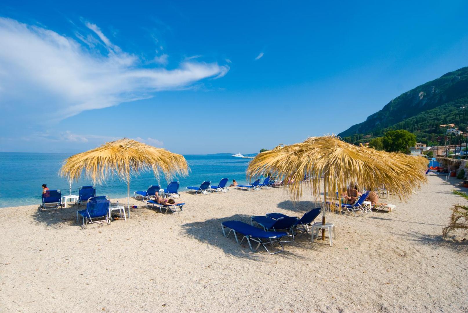 Potamaki Beach viesnīcas oļu pludmale ar ziliem sauļošanās krēsliem, salmu saulessargiem un skatu uz kalnaino Korfu piekrasti.