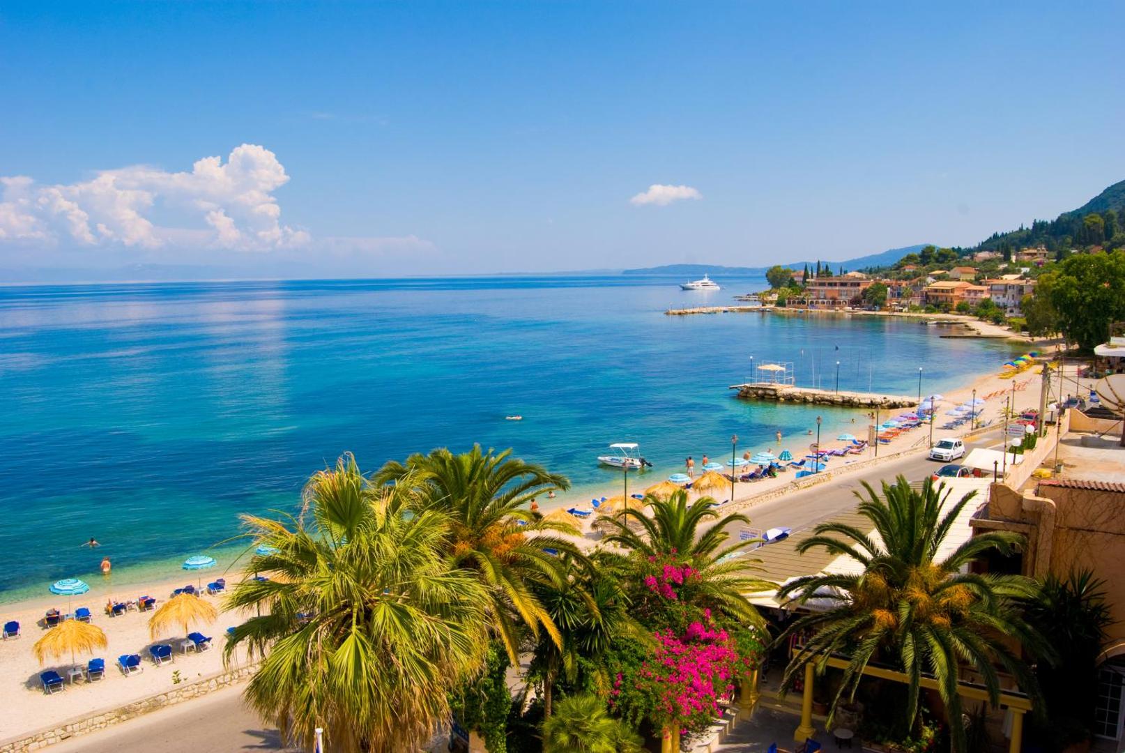 Skats uz Benitsesas pludmali Korfu salā no Potamaki Beach — tirkīza krāsas ūdens, saulessargi un palmas piekrastē