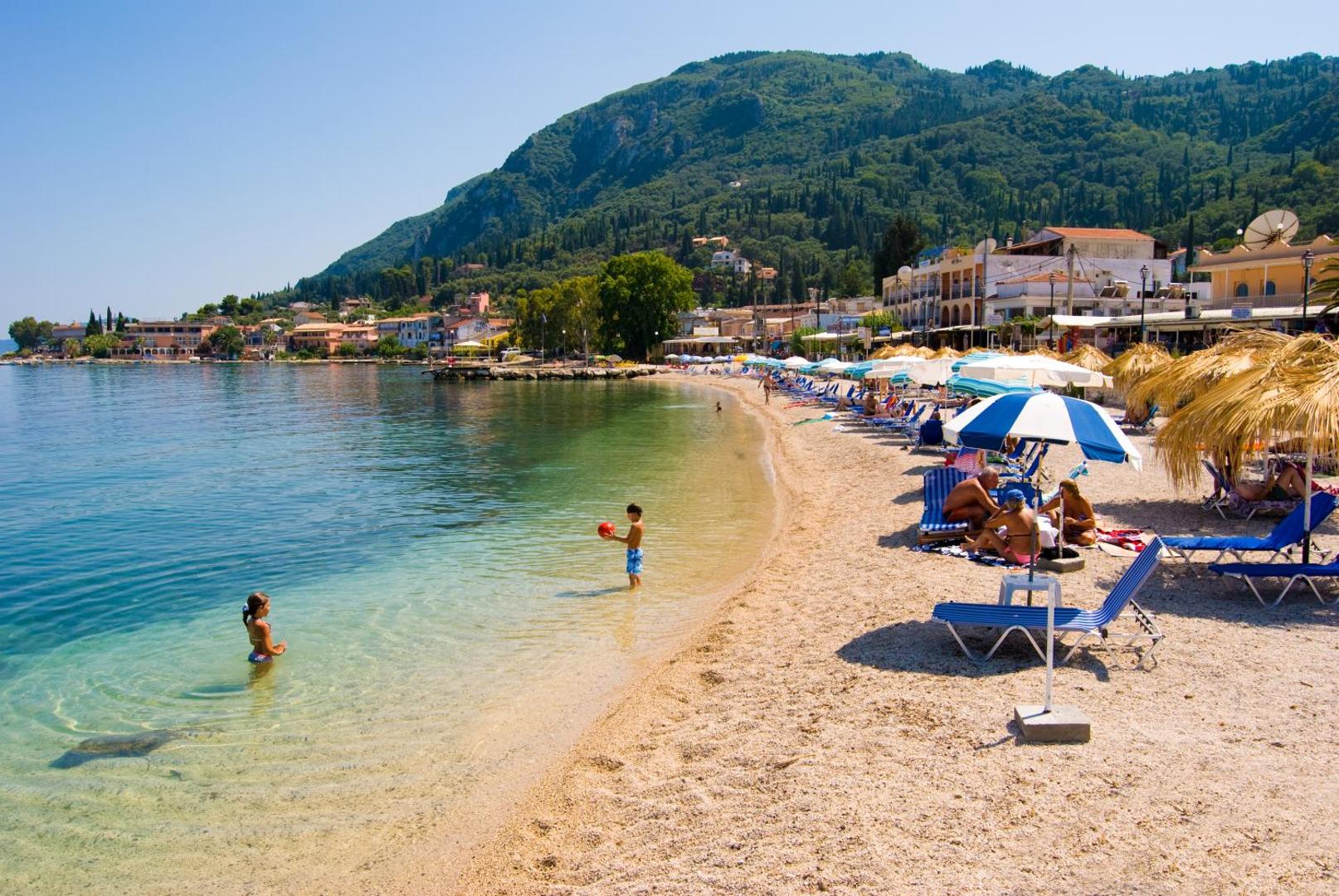 Bērni spēlējas seklajā ūdenī Potamaki Beach pludmalē Benitses ciematā, fonā redzama pilsētas apbūve un zaļi kalni.