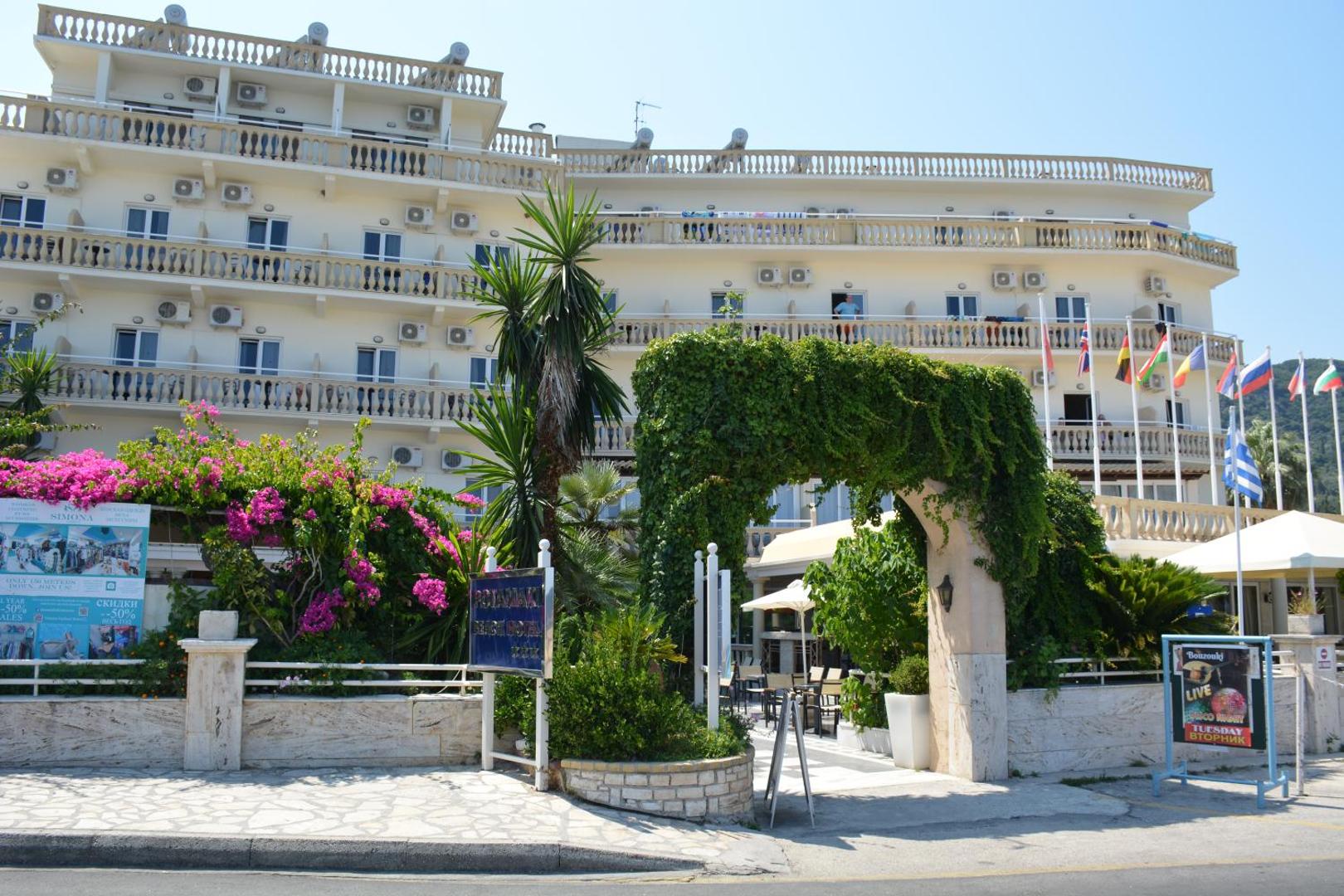 Potamaki Beach viesnīcas fasāde ar balkoniem, ziedošiem krūmiem un karogiem pie ieejas saulainā dienā