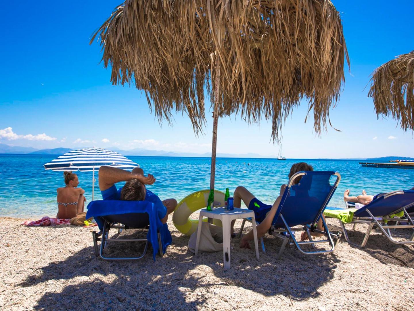 Viesi atpūšas Potamaki Beach viesnīcas pludmalē Korfu salā zem salmu saulessargiem pie dzidri zila Jonijas jūras ūdens.