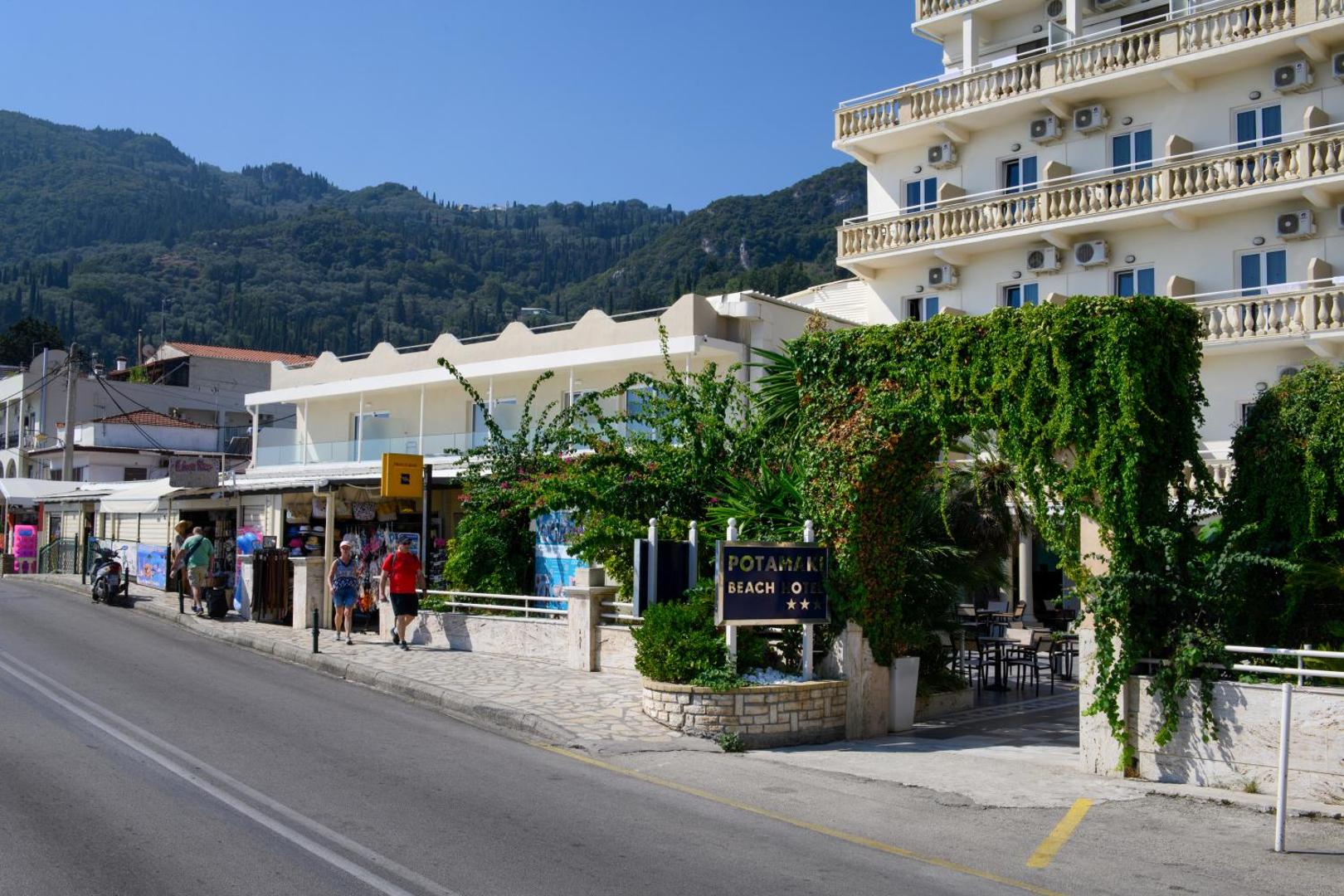Potamaki Beach viesnīcas ieeja no ielas puses ar norādi, zaļumiem klātu arku un kalnu ainavu fonā