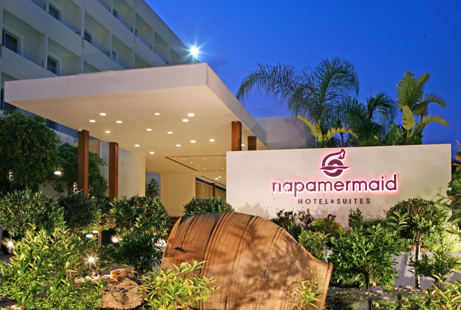 Napa Mermaid Hotel & Suites ieeja vakarā Ajanapā, Kiprā — izgaismots viesnīcas logo, dekoratīvi augi un moderna arhitektūra piekrastē.