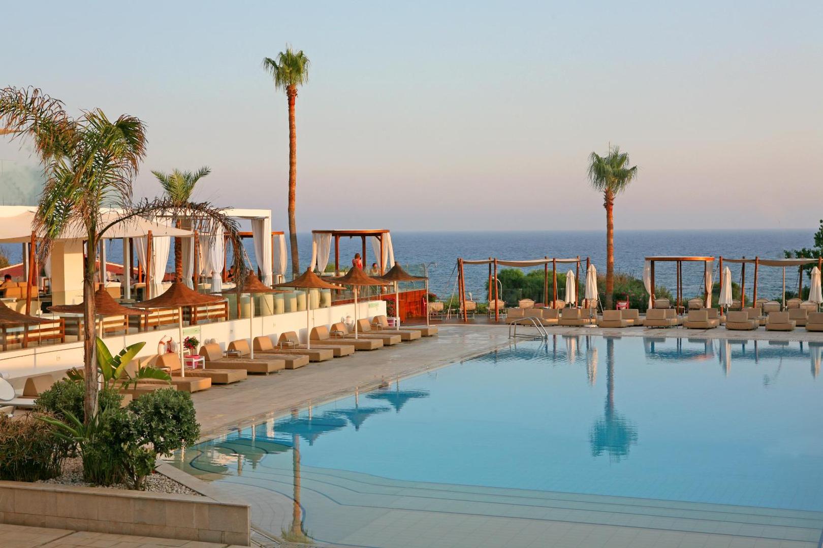 Napa Mermaid Hotel āra baseins ar sauļošanās krēsliem, saulessargiem un palmām piekrastē saulrieta laikā